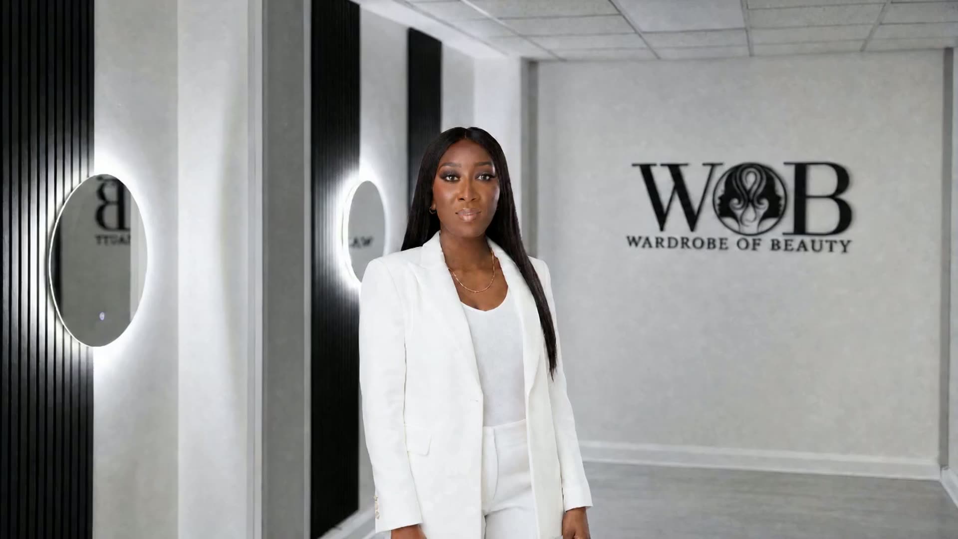 Bola Adepoju, founder of WOB Beauty, standing in the centre of the treatment room at 7 Hind Court with a warm open smile, the backlit round mirrors along the wall beside her and the black WOB Wardrobe of Beauty wall logo behind her.